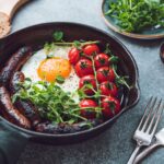 Breakfast time, fried egg with sausages and cherry tomatoes in a black iron pan, served microgreens.